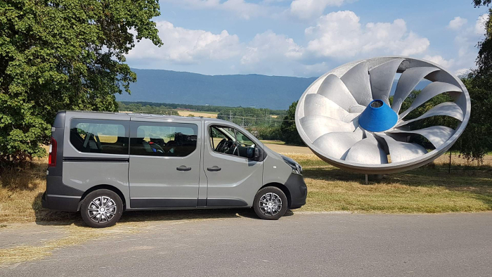 a photo of a grey minivan opel vivaro in a sunny countryside nex to a hydraulic turbine in Chancy/Geneva canton